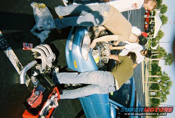 Several guys are gathered around the open hood of a blue car, working on the engine. Another car is visible in the background