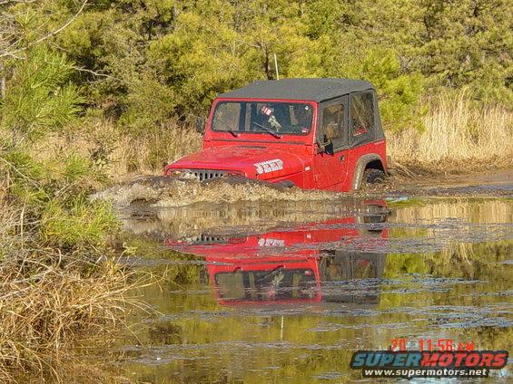Red Jeep Wrangler with soft top driving through muddy water, reflection visible