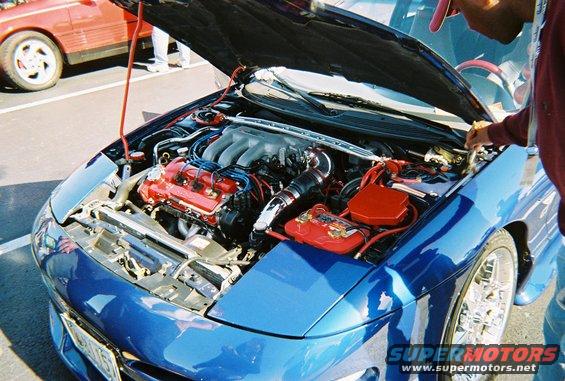 Blue car engine bay with V6 engine, red accents, and chrome wheels. Another red car is visible