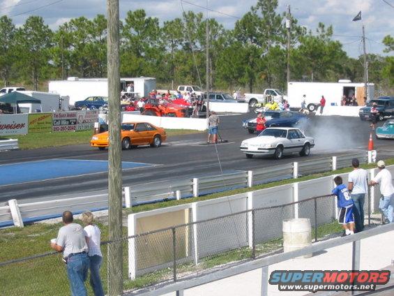 Two cars race on a drag strip, one kicking up smoke. Spectators watch from the side