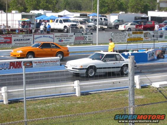 Orange and white Ford Mustangs on a drag strip, side view