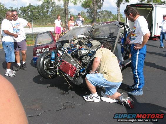 Front view of a severely damaged race car with exposed engine and "GULF COAST" text on the door