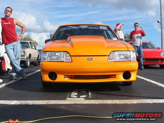 Front view of an orange Ford Mustang with a large aftermarket hood and racing decals on the windshield. Two men stand nearby
