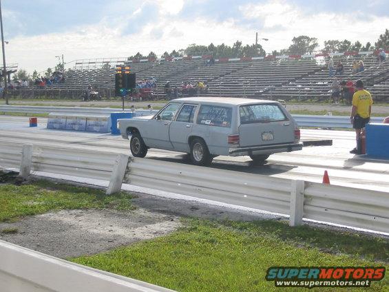 Gray station wagon at drag strip, driver in yellow shirt stands nearby