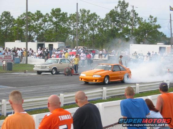 Drag strip with an orange Ford Mustang doing a burnout and a tan sedan in the background