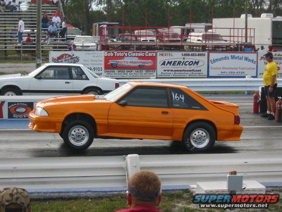 Orange Ford Mustang on drag strip, number 164 on door. White Ford sedan in background