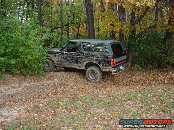Mud-covered black Ford Bronco parked on a dirt path in a wooded area