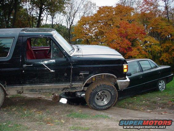 1988 Ford Bronco parked next to a dark sedan on a dirt path with fall foliage in the background