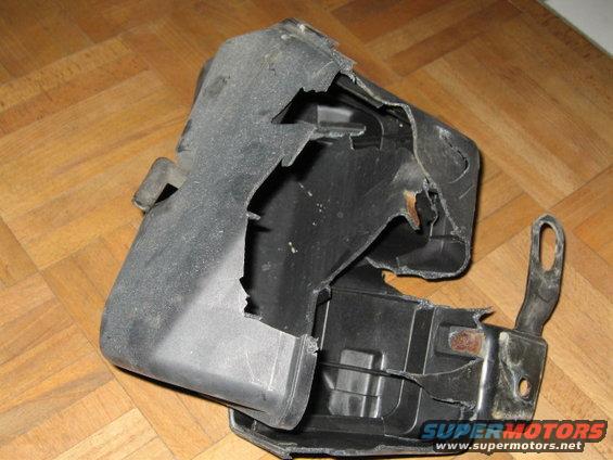 Damaged black plastic car part with metal bracket attached, lying on a wood floor