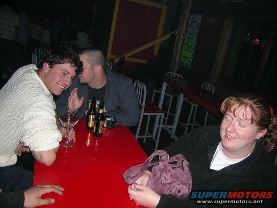 Guy whispering in another guy's ear at a red table with drinks. Woman in foreground with eyes closed