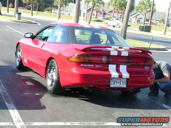 Red 1993 Ford Probe with white racing stripes, rear view, chrome wheels, spoiler, and a person inspecting the rear bumper