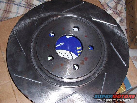 Slotted brake rotor with blue packaging visible through the center holes. Text on packaging reads 'parallel for anc'