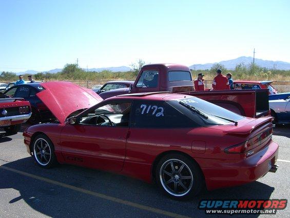 Red coupe with racing numbers on window, hood open, parked at an outdoor event