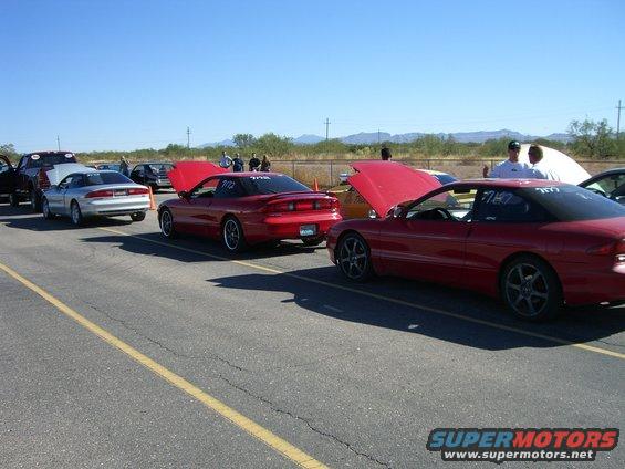 Red coupe with hood open, parked on asphalt with yellow line. Race numbers visible on door