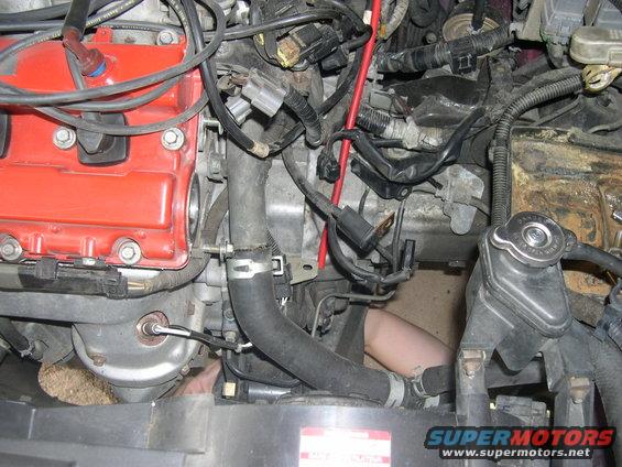 Close-up of car engine bay, red valve cover, hoses, wires, and a person's arm visible working on it