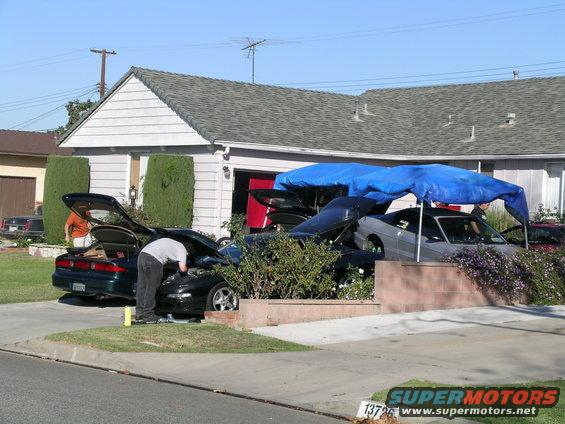 Two cars in a driveway, one dark green with trunk open, another black with hood open and a person working on it. A third car is partially covered by a blue tarp