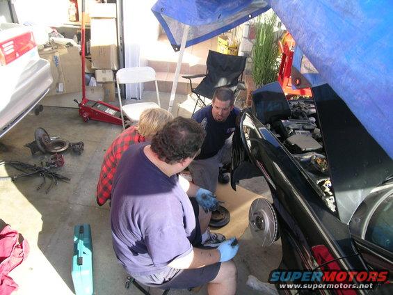 Three guys working on a black car with the hood up, one is holding a brake rotor