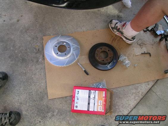 New slotted brake rotor next to old rusty one, with a box of hardware and a person's legs visible