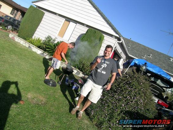 Man in dark t-shirt and shorts gives the finger while another man grills in the background