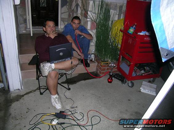 Two men sit outside with hookahs and a laptop, next to a red toolbox