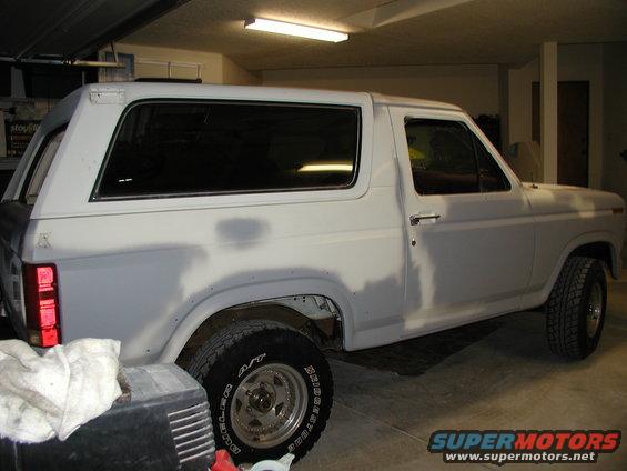 White SUV, possibly a Ford Bronco, in a garage. It has primer on the body panels and appears to be undergoing restoration