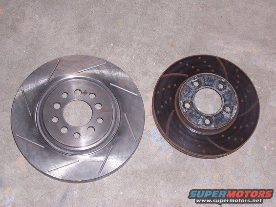 Two brake rotors on a concrete surface; one is new with slots, the other is old and rusted with drilled holes