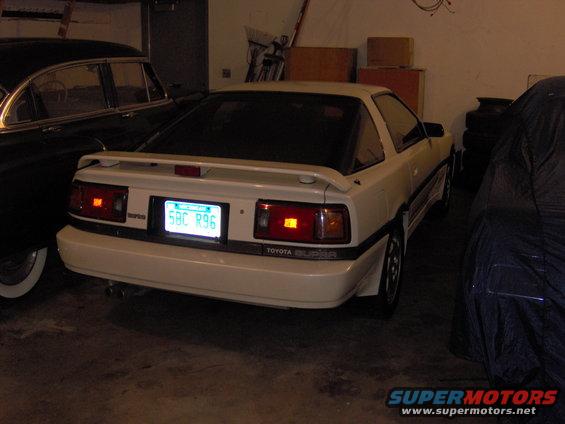 White Toyota Supra parked in garage next to a dark car and a covered vehicle. Rear view shows taillights and license plate