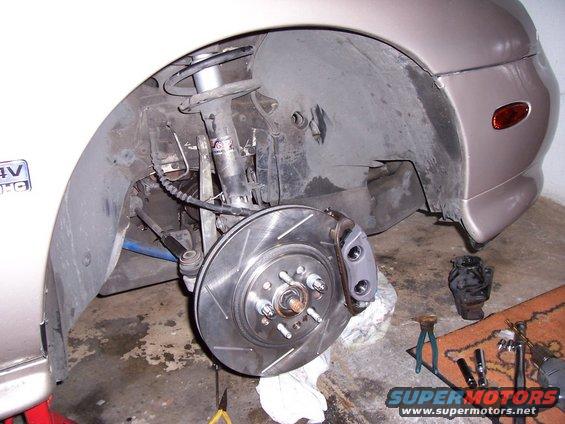 Close-up of a car's front wheel well showing the brake rotor and caliper. Tools are on the floor
