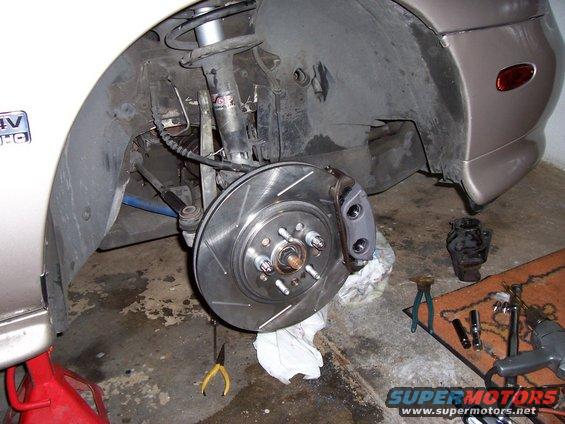 Car brake rotor and caliper, tan car body, red jack stand, tools on floor