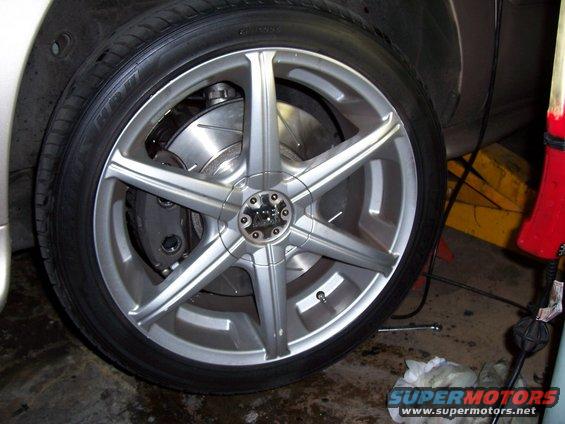 Silver alloy wheel with six spokes, showing brake caliper and rotor. Tire is black