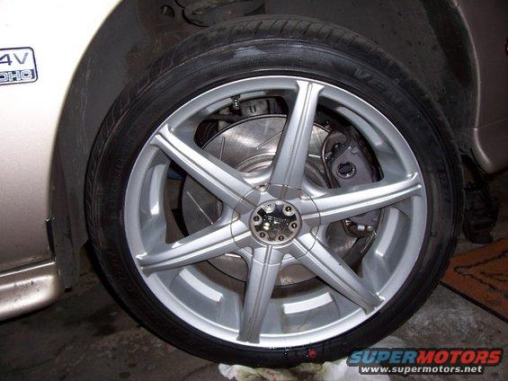 Close-up of a silver 5-spoke aftermarket wheel with a drilled brake rotor and caliper. A '4V DOHC' badge is visible on the car's body