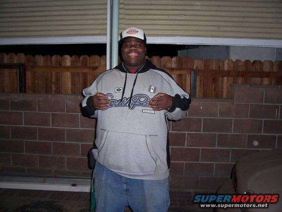 Man wearing a gray hoodie with 'South Pole' logo and a white baseball cap stands in front of a brick wall and wooden fence