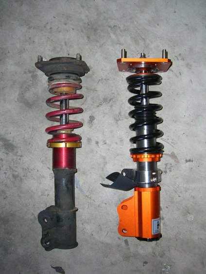 Two car suspension struts, one black and red, the other orange and black, sit side-by-side on a concrete floor