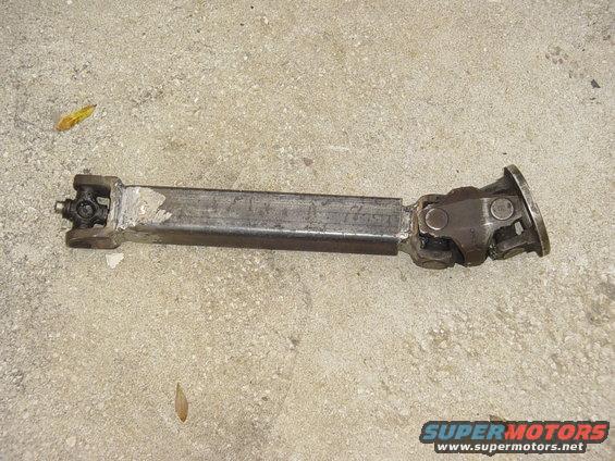 Driveshaft with universal joints on a concrete surface. The metal shaft shows signs of welding and wear