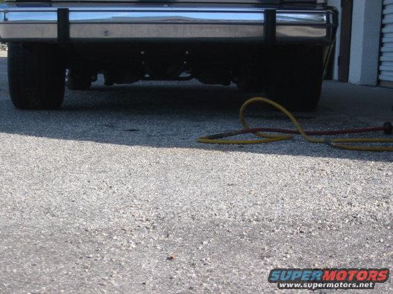 Car's chrome bumper with black protective strips, rear tires, and a yellow and red hose on the ground