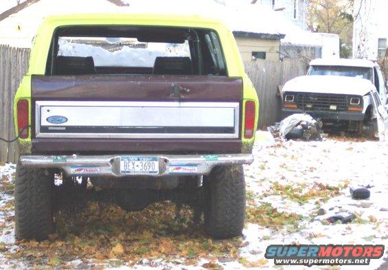 Rear view of a yellow and brown Ford Bronco with a New York license plate BEX-3690, another white Ford truck in the background