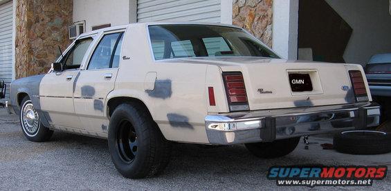 Rear view of a beige Ford sedan with primer patches on the body, black wheels, and a license plate that reads GMN