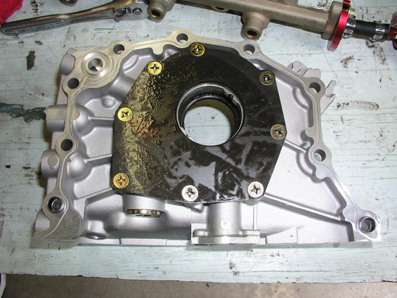 Engine oil pump housing with black gasket and gold screws, resting on a wooden surface. Tools are visible in the background
