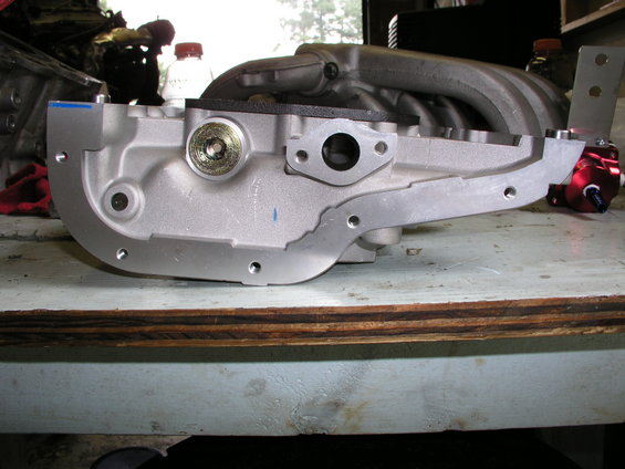 Aluminum engine intake manifold with red fuel pressure regulator attached, resting on a workbench
