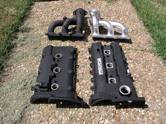 Two engine valve covers and two exhaust manifolds on gravel. One valve cover has "Mazda" text