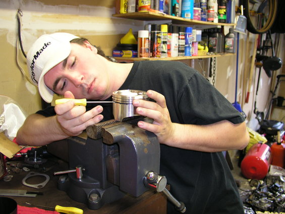 Man in white cap with yellow screwdriver works on piston in vise