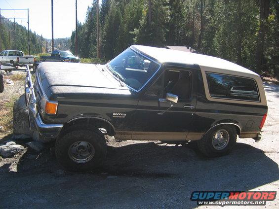 1990 Ford Bronco parked on dirt road with trees in background. Two other trucks visible