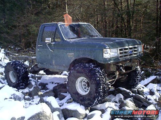 Green Ford F-350 pickup truck with large off-road tires, parked on rocks in the snow