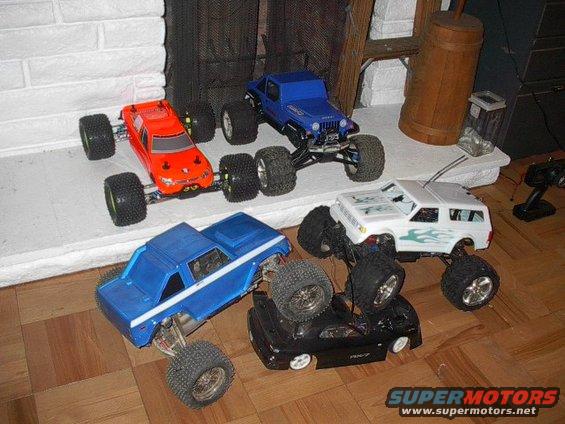 Several remote control monster trucks are arranged on a wooden floor and a white hearth
