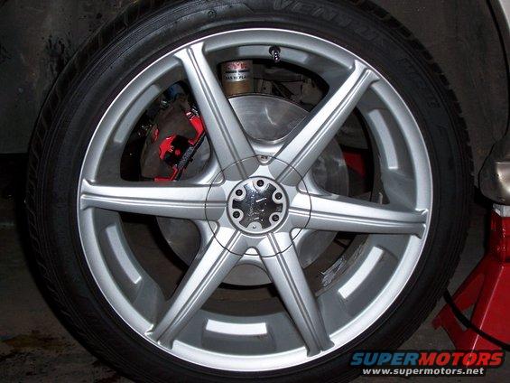 Close-up of a silver multi-spoke car wheel with red brake caliper visible behind it. A jack stand is on the right