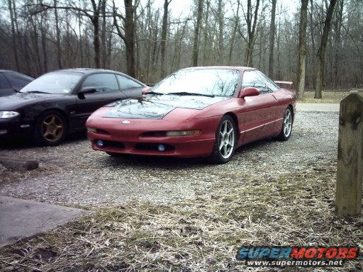 Red 1995 Ford Probe with a black hood parked next to a dark car in a gravel lot with trees in the background
