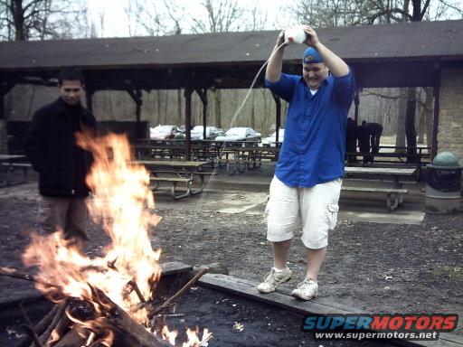Two men by a campfire, one pouring liquid from a bottle