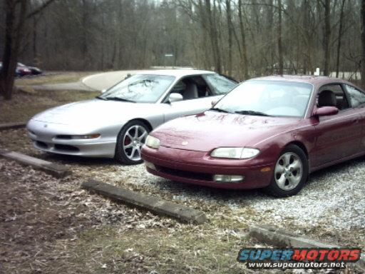 Maroon and silver coupes parked on gravel lot with trees in background. "SUPERMOTORS" logo visible
