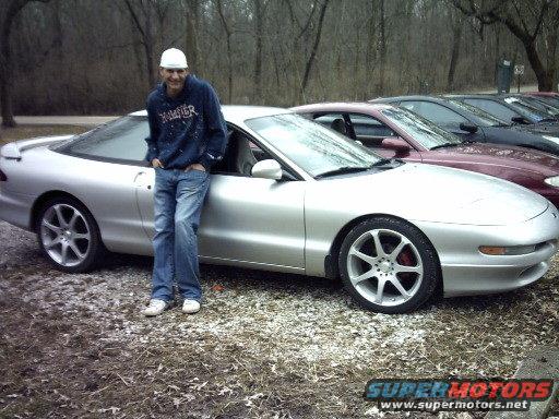 Man in blue hoodie and jeans leaning on a silver 1995 Ford Probe; other cars in background
