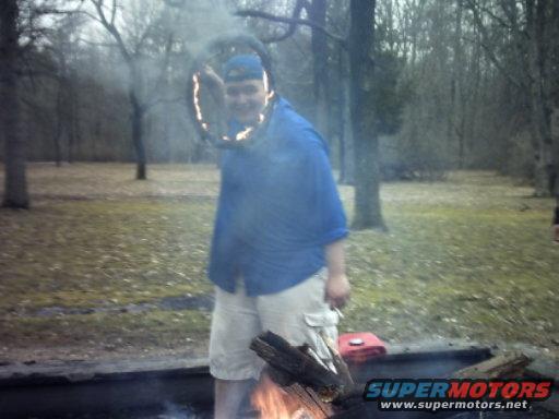Person in blue shirt and khaki shorts holds flaming tire rim in wooded area with campfire